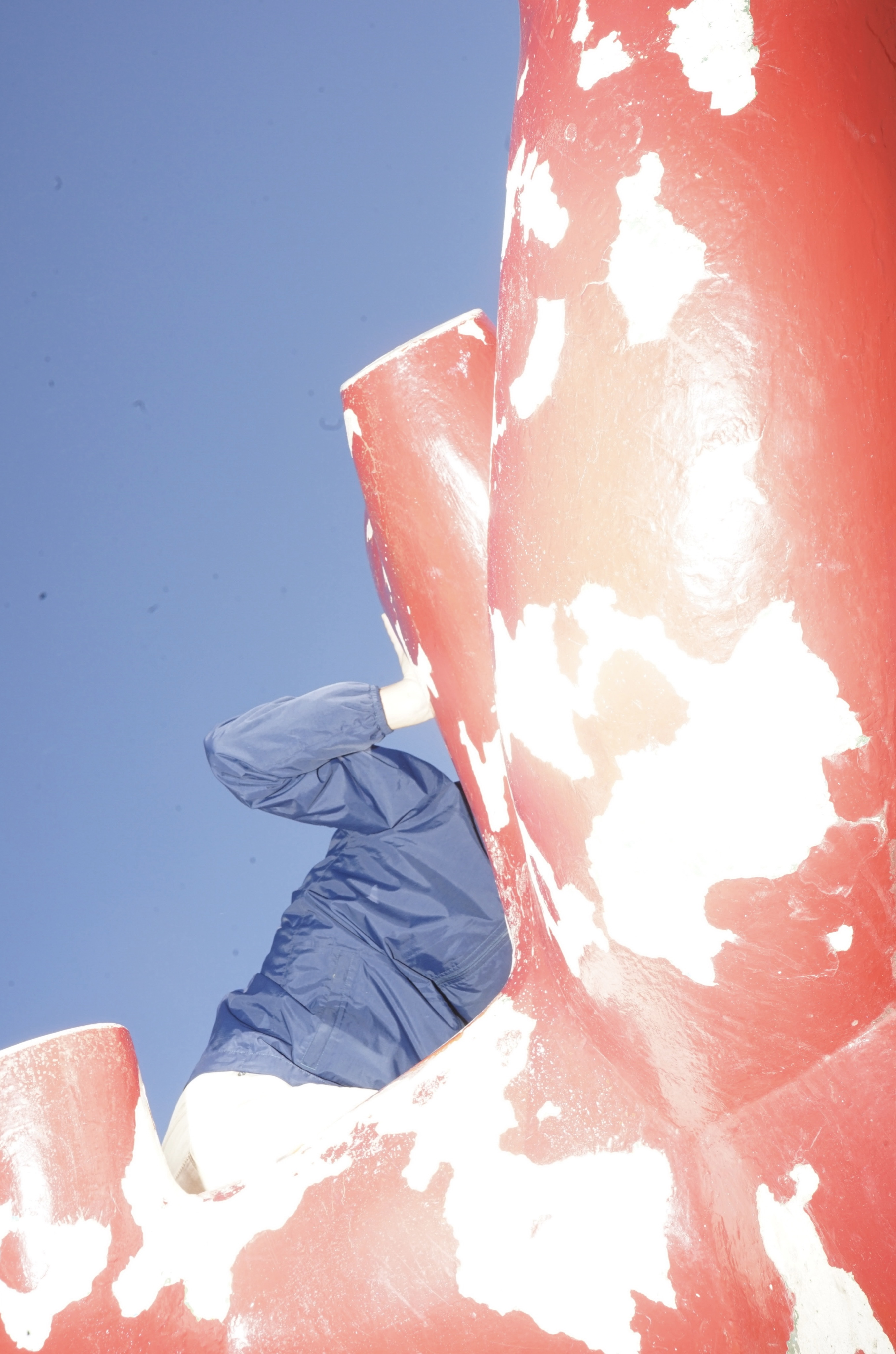 A low-angle shot shows a person in a blue windbreaker climbing behind a large, chipped red sculpture under a bright blue sky.