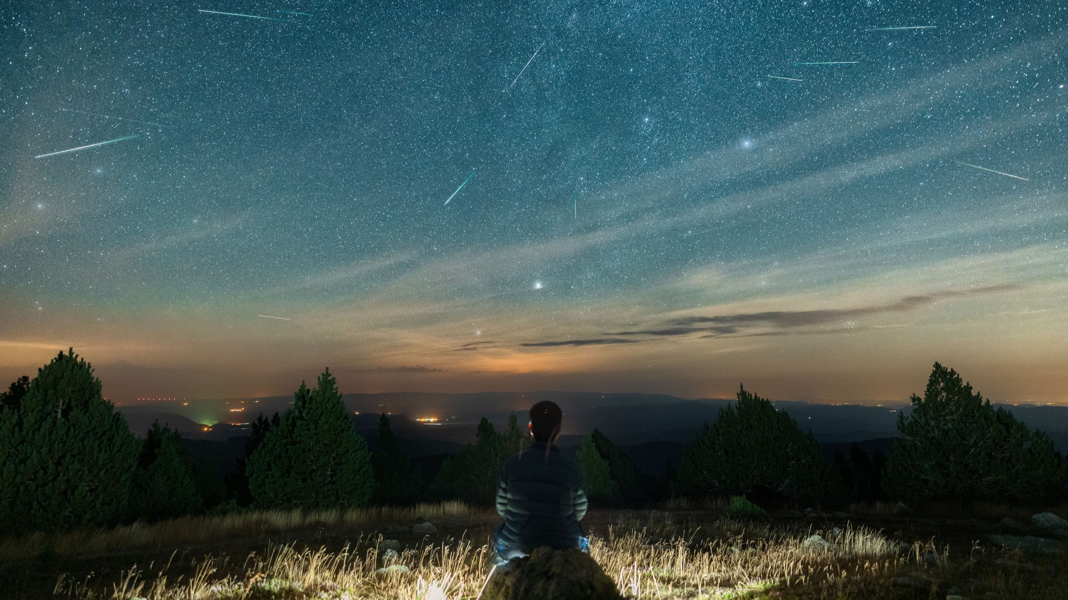 a man sits on a rock and looks at a sky filled with long streaks of light - perseid meteors.