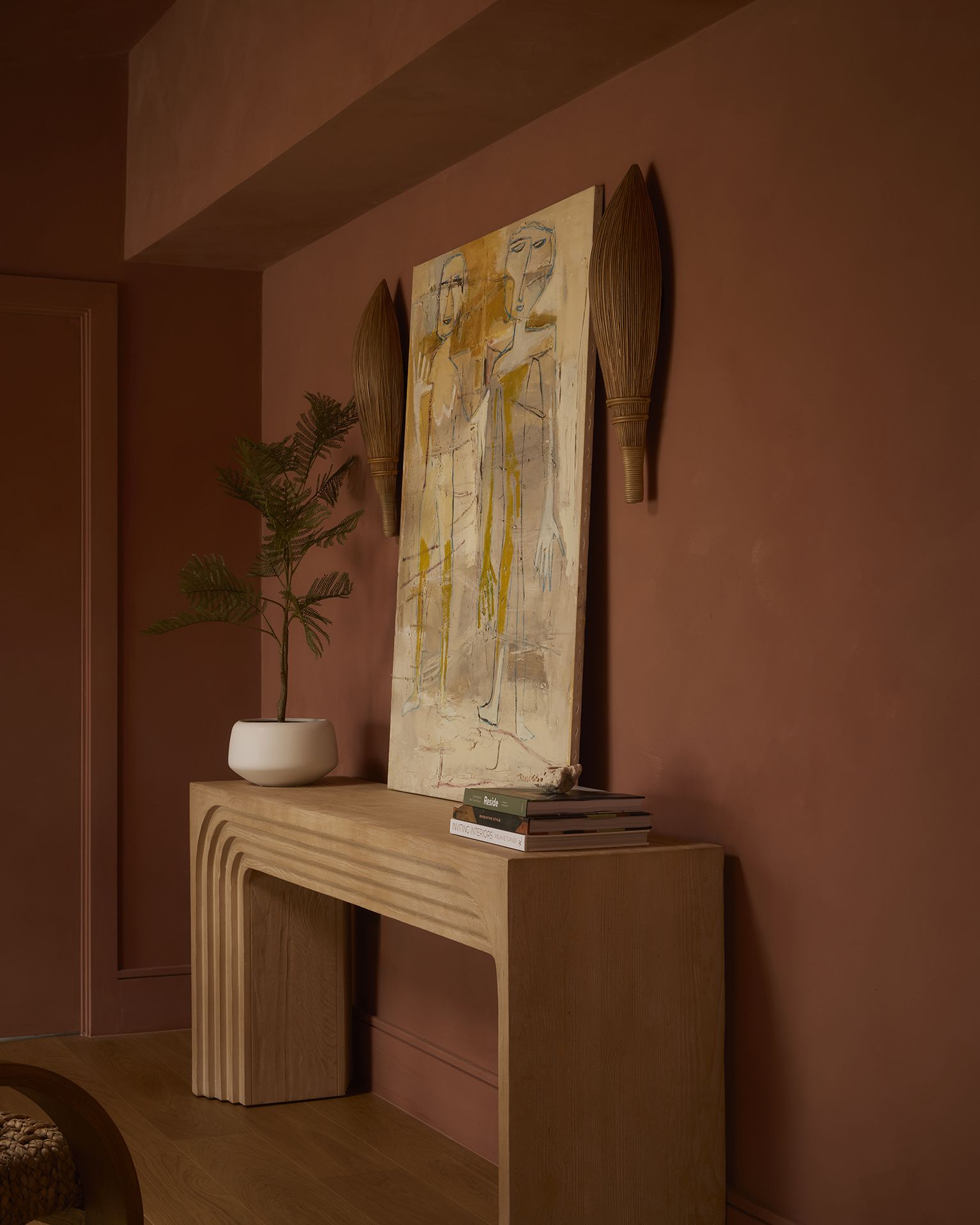 A bench-shaped table in textural wood stands against a terracotta wall topped by an abstract canvas, a vase, and books.