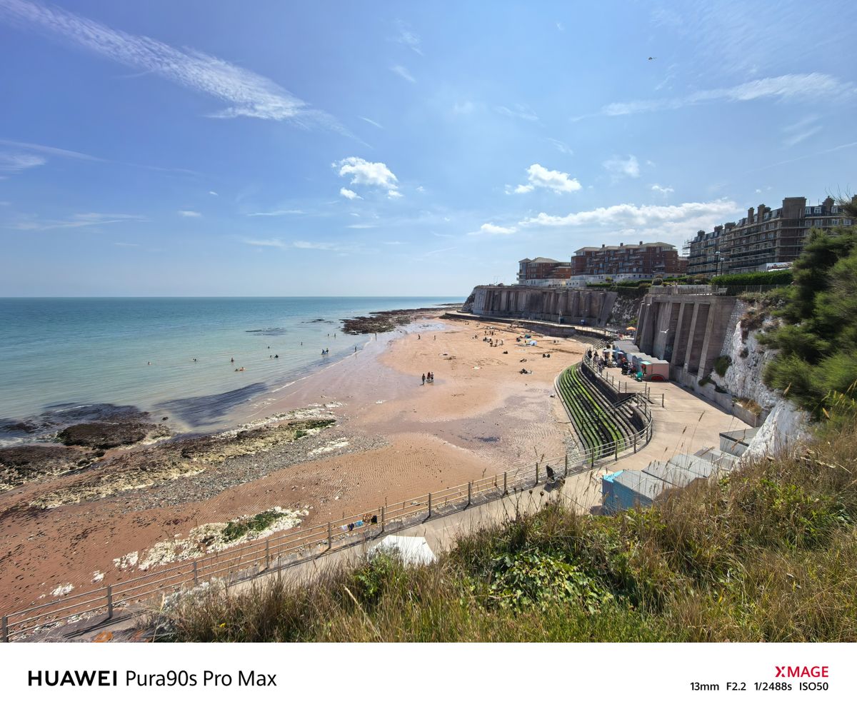 Wide coastal view of a sandy beach beneath chalk cliffs and seafront buildings on a bright sunny day.
