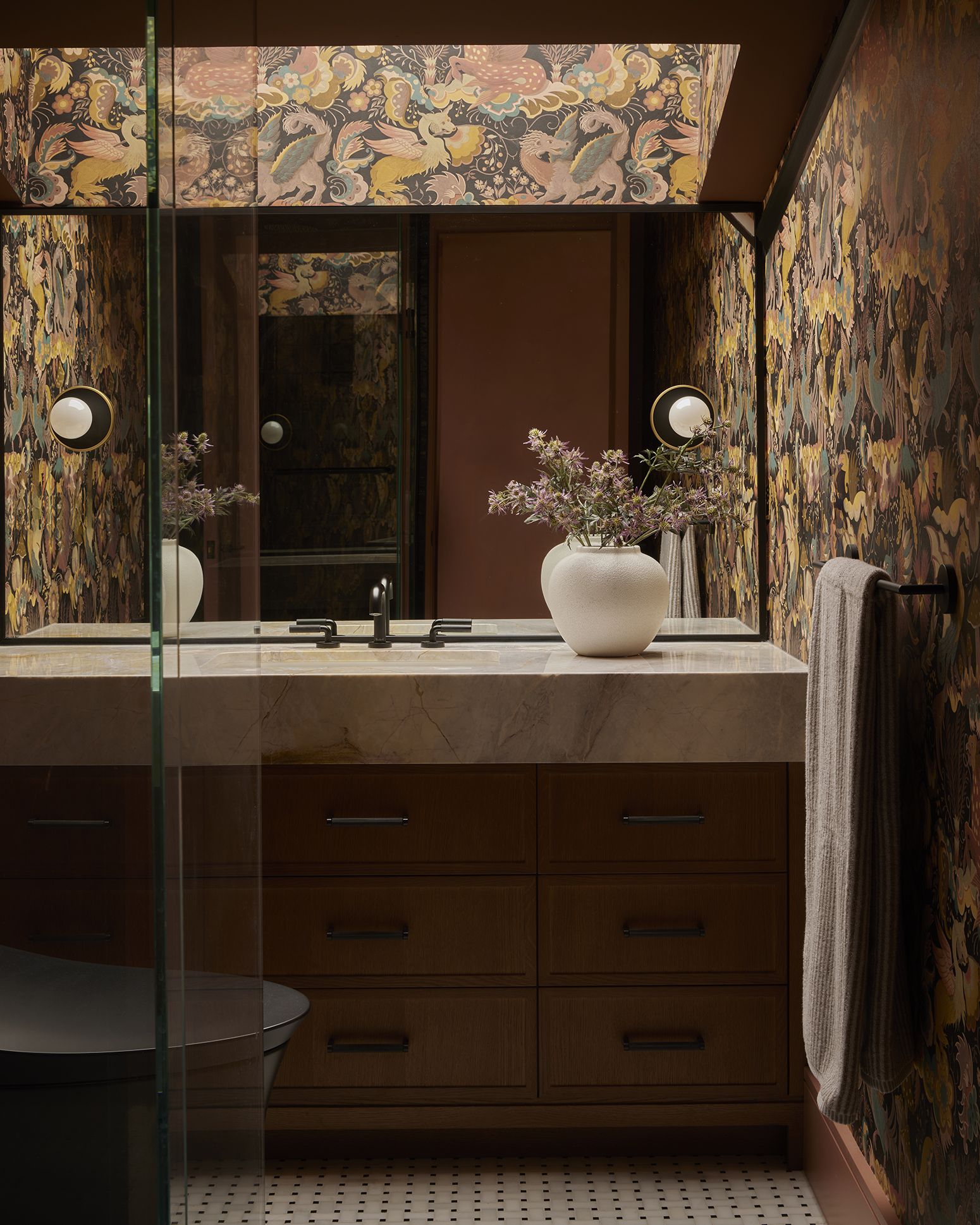 A maximalist bathroom with patterned painted walls, a wood and marble basin with an integrated chest of drawers, a sculptural vase, tiled floors, and round lighting.