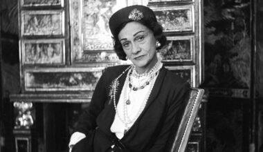 Seated on the mirrored staircase at 31 Rue Cambon, Chanel wears a tweed skirt suit, hat, and cat-eye glasses as she watches h
