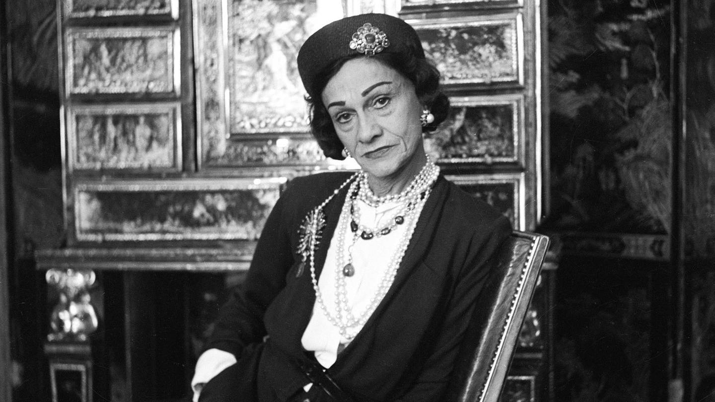Seated on the mirrored staircase at 31 Rue Cambon, Chanel wears a tweed skirt suit, hat, and cat-eye glasses as she watches h
