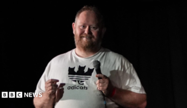 A man in a white t-shirt stands facing the camera on a stage before a black background and smiles towards the crowd in front of him. He holds a microphone in his left hand. he has short brown hair and a gingery- brown beard.