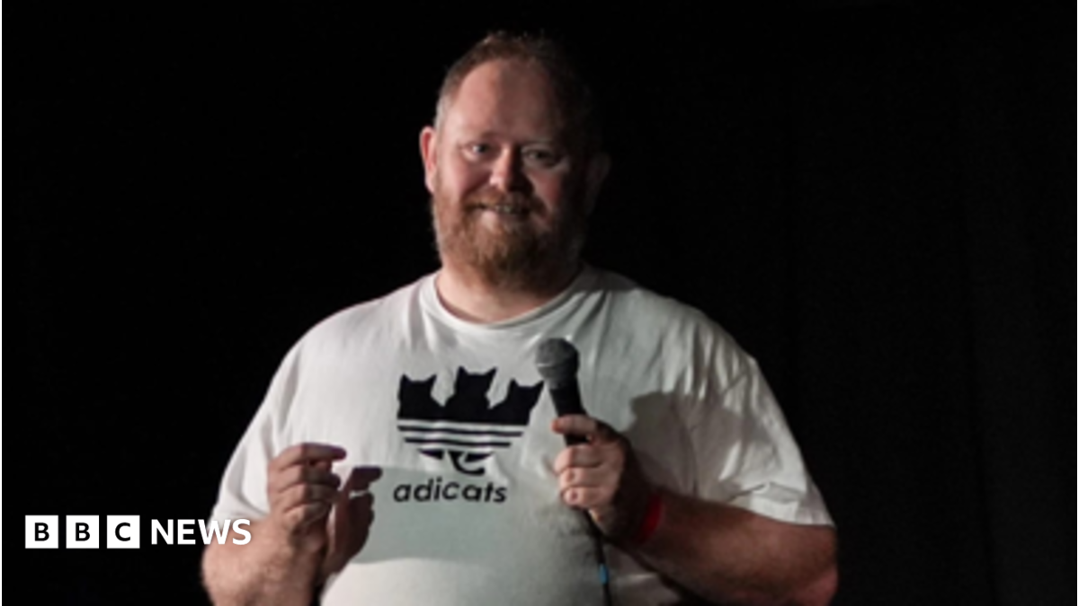 A man in a white t-shirt stands facing the camera on a stage before a black background and smiles towards the crowd in front of him. He holds a microphone in his left hand. he has short brown hair and a gingery- brown beard.