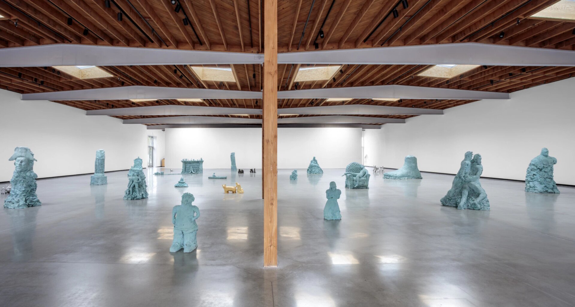 Wide gallery view with various abstract blue sculptures on polished concrete floor and wooden ceiling beams.