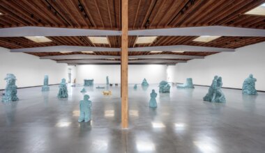 Wide gallery view with various abstract blue sculptures on polished concrete floor and wooden ceiling beams.