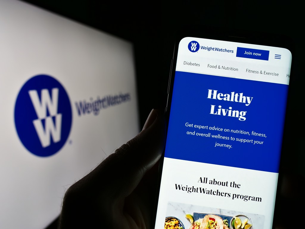 A person holding a phone displaying the Weight Watchers website with "Healthy Living" and program details, in front of a blurred Weight Watchers logo