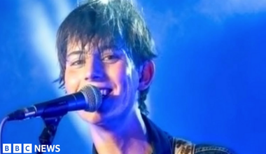 A close-up image of Kit Bottomley with short brown hair, who is smiling and singing into a microphone against a blue backdrop.