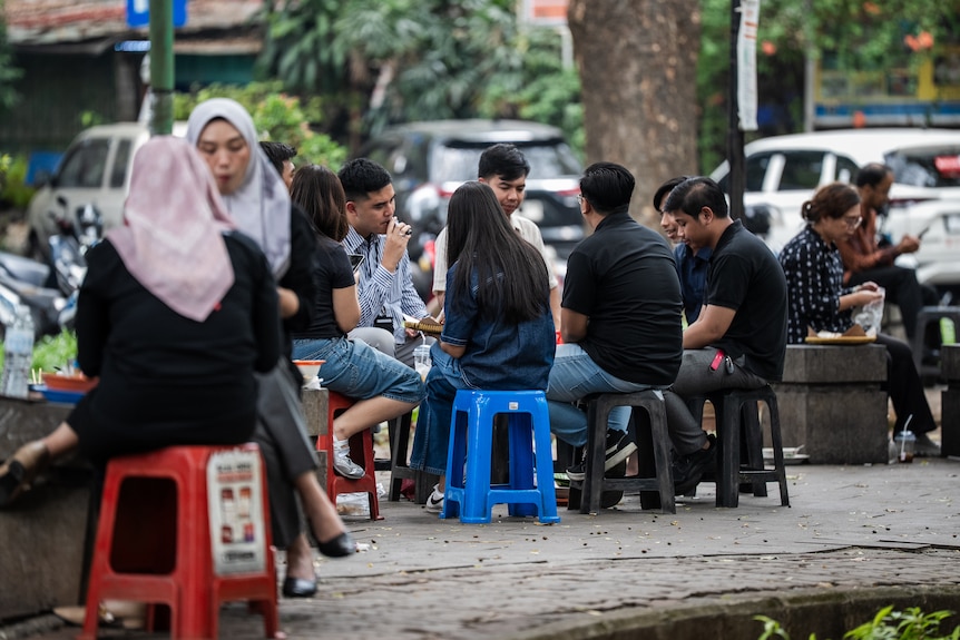 People sit outdoors on plastic stools on a footpath, eating street food and talking.