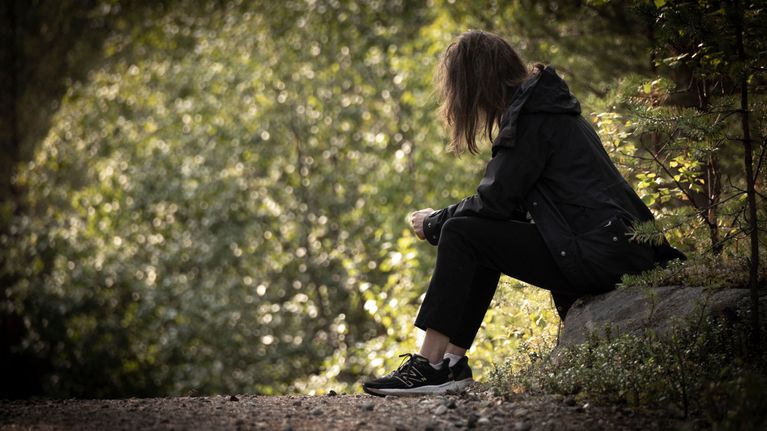 Woman in the woods sitting on a rock, seen from the side, her shoulder length hair is concealing her face.