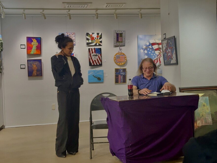 One person is sitting behind a desk while a second person stands next to the desk. Both of them are in an art gallery.