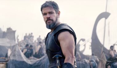 A bearded man in medieval armor stands in front of a busy camp with tents and people, holding a sword at his side, with ships in the background.