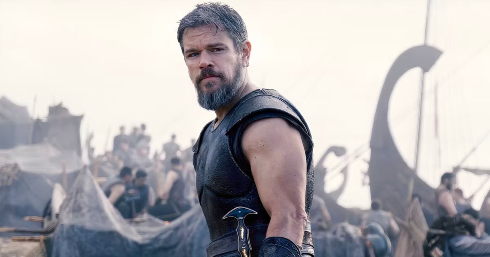 A bearded man in medieval armor stands in front of a busy camp with tents and people, holding a sword at his side, with ships in the background.