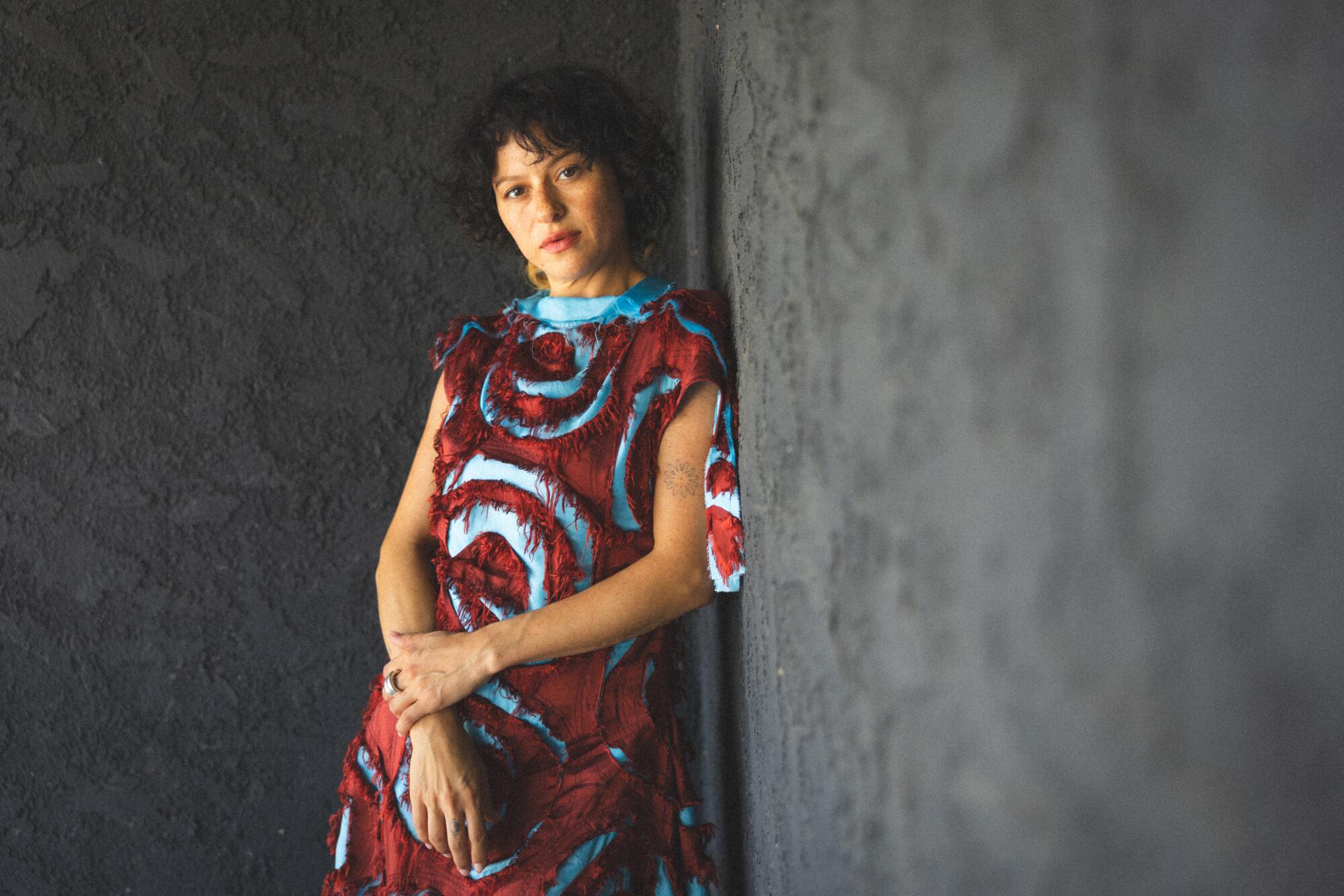 A woman leans against a wall in a maroon and blue dress.