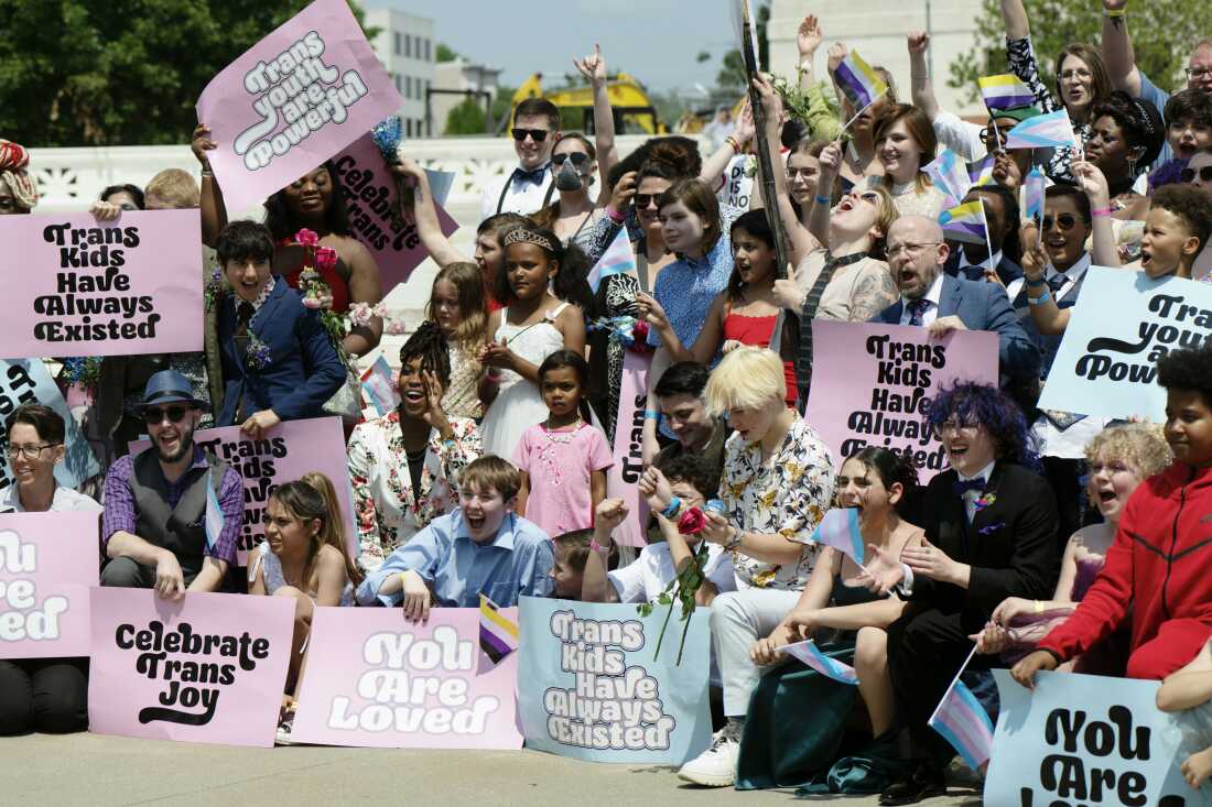 A boisterous crowd of mainly young people fills the photo frame. Printed signs say "Trans kids have always existed" and "Celebrate trans joy," among other slogans.