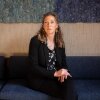 Noella Sudbury is the founder of Rasa Legal, a non-profit that uses technology to help people expunge their criminal records. She is sitting on a couch with her hands in her lap. Sudbury has long, curly hair and she is wearing a dark suit and a dark floral top.