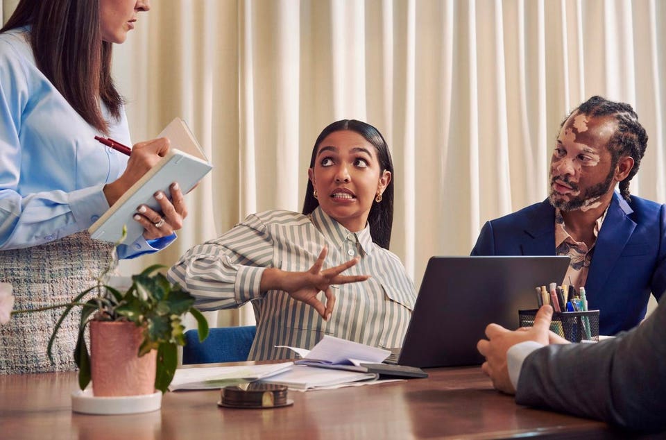 A Deaf Latina Puerto Rican woman communicates in American Sign Language to a caucasian woman with long straight brown hair wearing business attire standing next to her while seated at a conference room table with two colleagues.