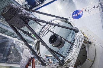 The Roman space telescope is positioned in the foreground against a backdrop of glowing purple nebulae and numerous galaxies. The telescope features a dark, angular structure with a large central mirror and several rectangular components. The scene conveys a sense of depth and vastness in space.