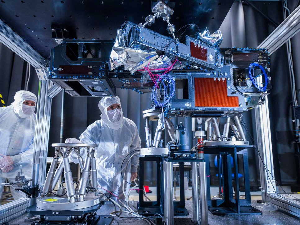 A laboratory setting with two people wearing protective suits and surrounded by complex technical equipment.