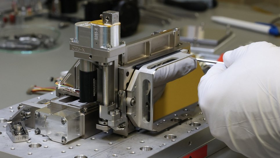 Close-up of a precision alignment mechanism on a workbench. The device features several metal components with visible screws, guides, and a yellow cover. A person wearing white gloves is holding a tool and working on one of the screws. Additional tools and parts are out of focus in the background.