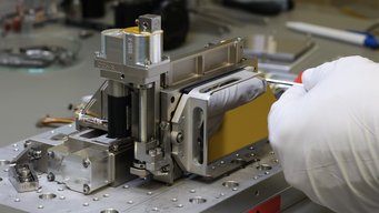 One of six flight models of the Precision Alignment Mechanisms (PAMs) for the Coronagraph Instrument (CGI), a camera aboard the Nancy Grace Roman Space Telescope. The PAMs position and stabilize the optical elements of the CGI during observations. Close-up of a precision alignment mechanism on a workbench. The device features several metal components with visible screws, guides, and a yellow cover. A person wearing white gloves is holding a tool and working on one of the screws. Additional tools and parts are out of focus in the background.