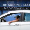 An electric display sign along a street says "THE NATIONAL DEBT" in white capital letters against a blue background. Underneath the words is the U.S. government's debt, which in this electronic display is $40,047,520,904,160. A light-colored car driven by a man passes by on the street.