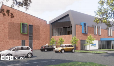 The building is low level and has brown  brick coloured walls, with two wings in the image. There is a car park with cars in the foreground.