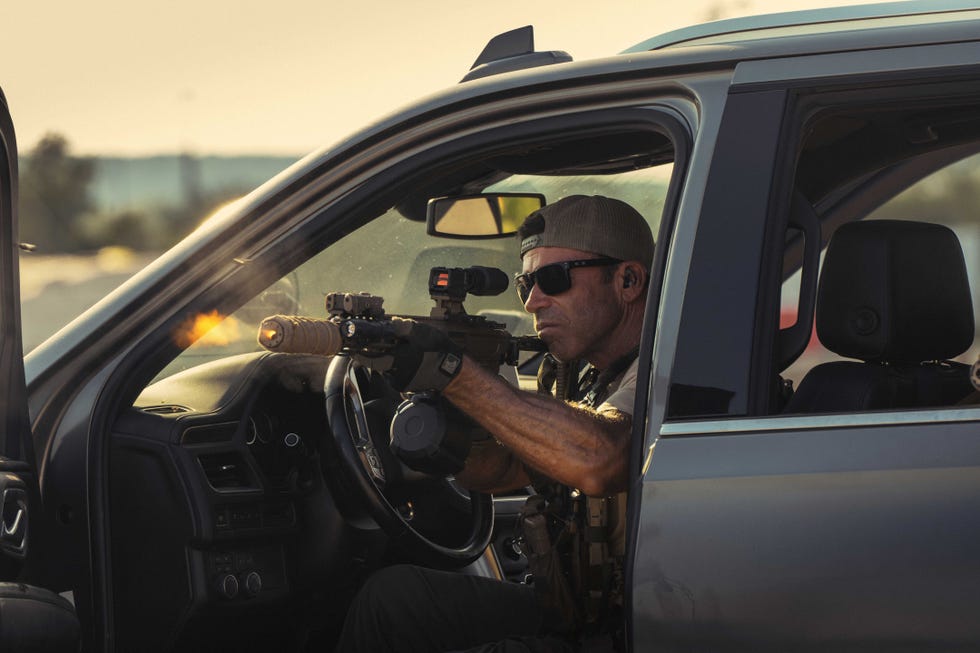 A man in tactical gear aiming a firearm from the driver's seat of a vehicle.