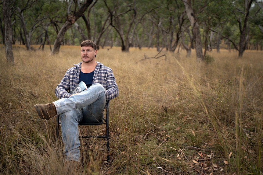 Luke Bateman, wearing a checked shirt and jeans, sits on a chair in long grass with trees in the background.