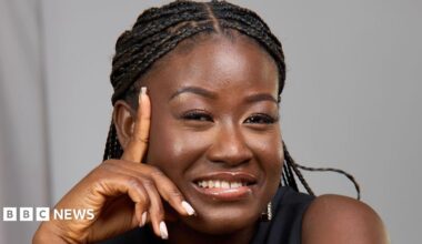 A woman with braids is smiling and leaning her face on her hand. She is wearing a black top and is photographed in front of a grey background.