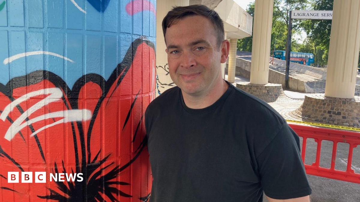 A man with short brown hair and a black T-shirt is smiling while standing next to a pillar painted blue with a red poppy design.