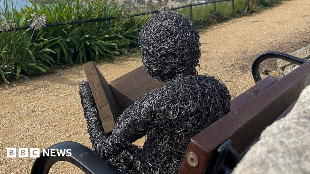 A metal sculpture of a human figure reading a book on a wooden park bench