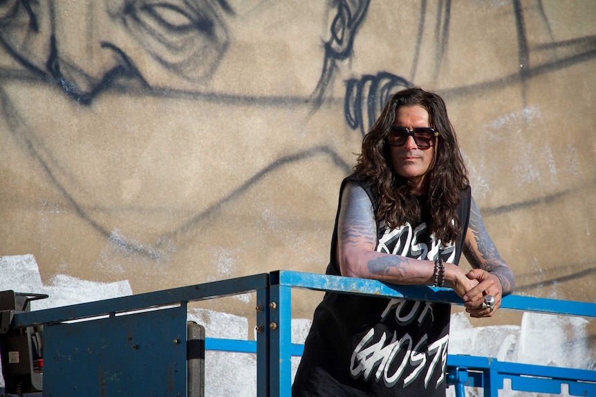 Artist Stormie Mills poses in front of his work in Kalgoorlie. He has long hair and is wearing sunglasses