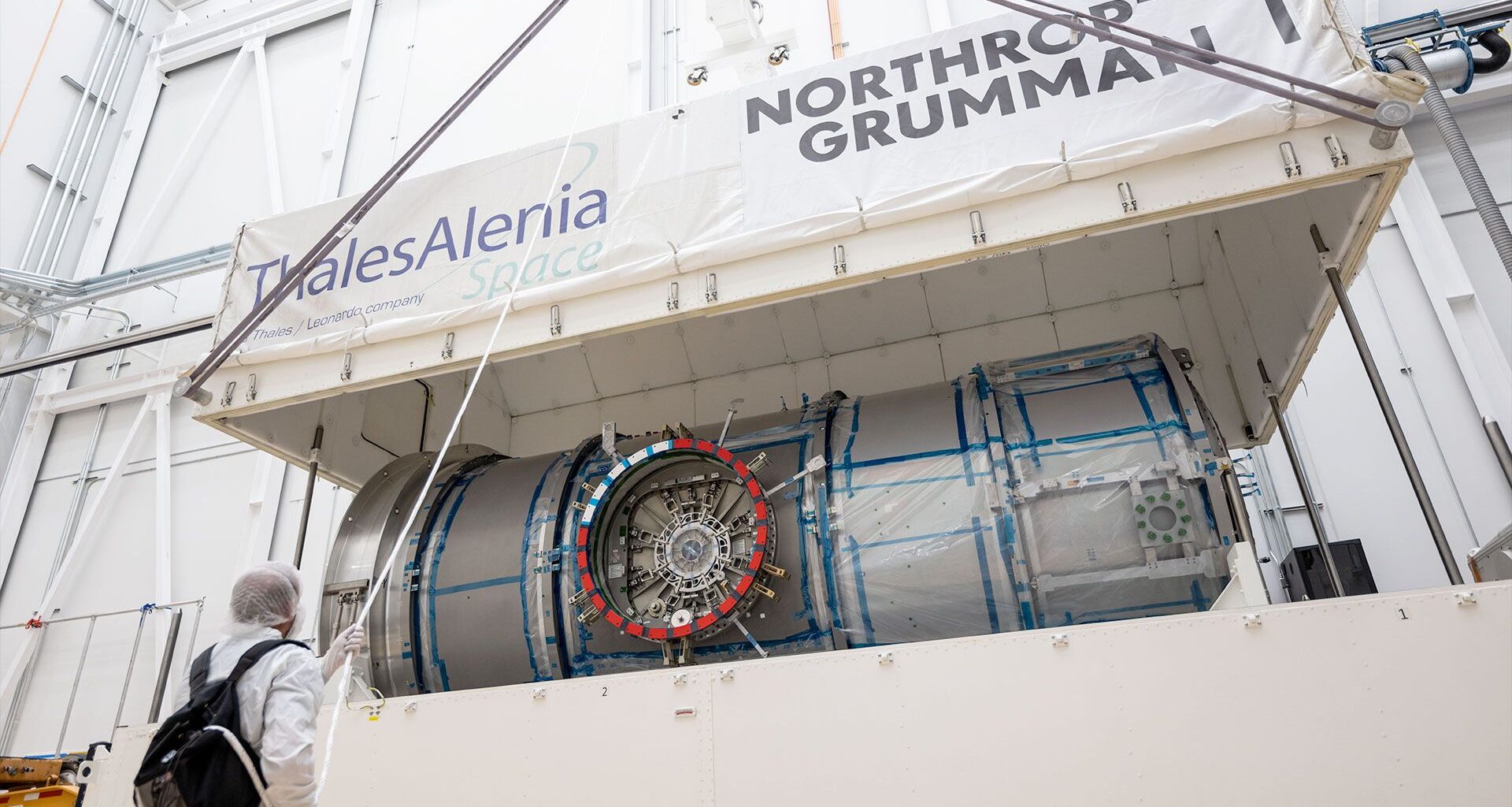 a white top half of a large shipping container is lifted by crane, exposing a metal cylindrical module inside.
