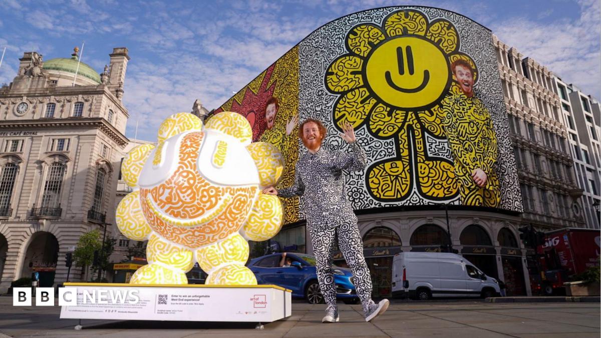 Mr Doodle stands beside one of his sculptures in Piccadilly Circus. The installation features his signature black-and-white doodle artwork with bright orange and yellow colours, while an image of the artist appears on the famous Piccadilly Circus screen behind him