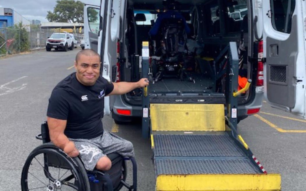 Barney Koneferenisi is in his wheel chair. You can see that he doesn't have legs below the knee. He is pointing towards the back of a wheelchair accessible van which has it's tail lift down.