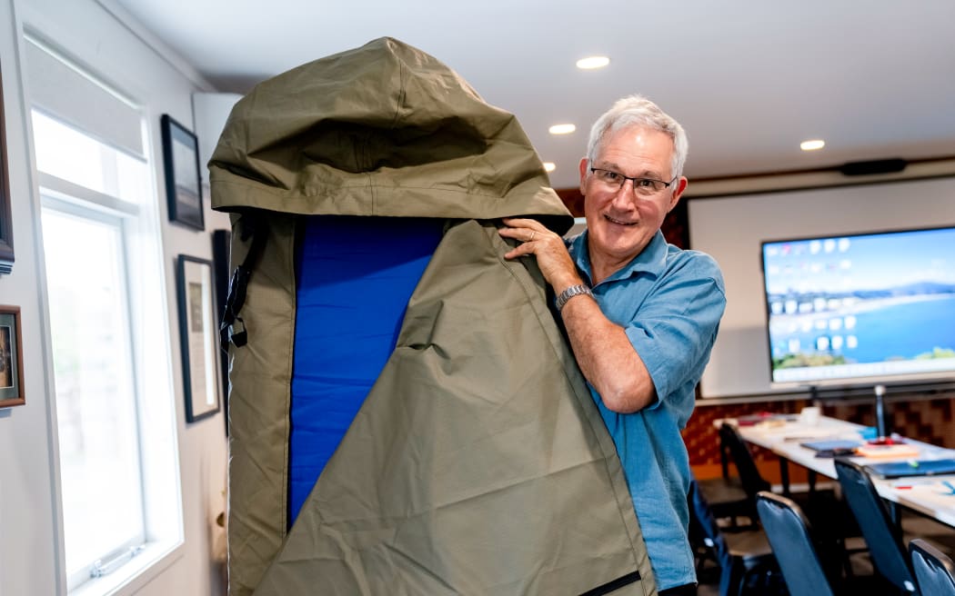 Man in glasses stand holding up canvas sleeping bag with in built mattress
