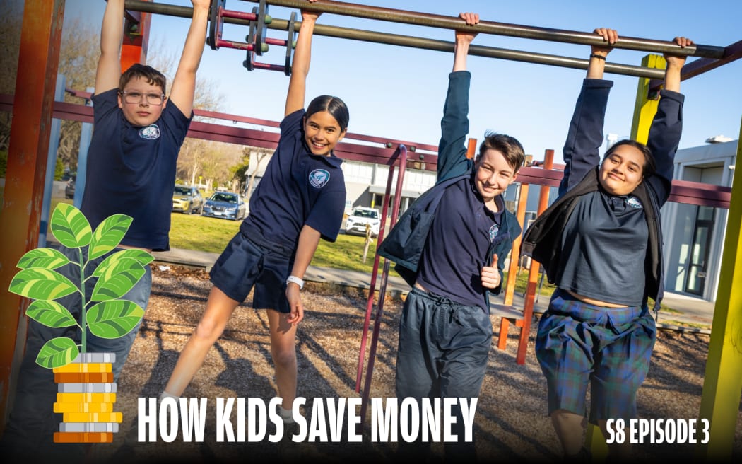 Four primary school students in navy uniforms hang from monkey bars on a sunny playground, grinning at the camera. One gives a thumbs up. A wood chip ground surface, parked cars, and school buildings are visible in the background.
