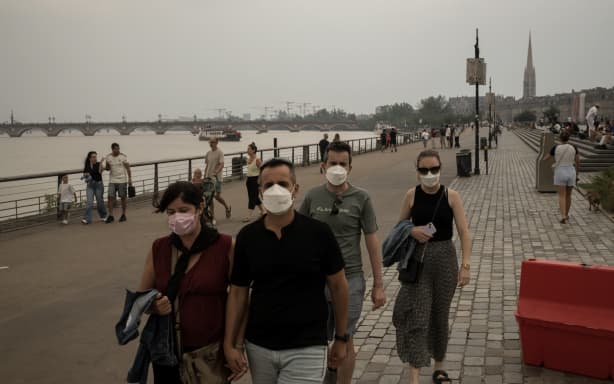People wearing masks to protect themselves from smoke walk along the river in central Bordeaux, southwestern France on July 26, 2026. France fought an "unpredictable" wildfire close to Bordeaux on July 26, 2026 while Spain warned of "difficult hours" in its own fiery battle, with evacuations from danger zones in both countries mounting to well over 300,000 people. Scientists have long warned that climate change caused by human use of fossil fuels will make periods of drought more intense and longer-lasting, creating the ideal conditions for wildfires. (Photo by Ed JONES / AFP)