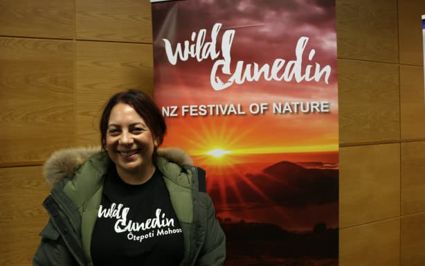 Amanda is wearing a black 'Wild Dunedin' t-shirt underneath a khaki green hooded jacket. She is smiling and standing in front of a Wild Dunedin: NZ Festival of Nature banner featuring a bright sunset.