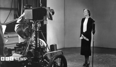 A black and white image from 1936 - a smartly dressed lady stands in front a microphone and a television camera