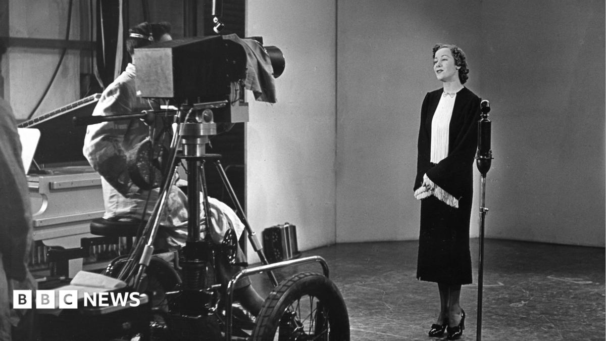 A black and white image from 1936 - a smartly dressed lady stands in front a microphone and a television camera