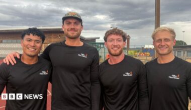 Four men standing on a running track. They are all wearing long-sleeve black tops with a white and orange Miles That Matter logo on the chest. The man on the left has black curly hair. The man next to him has brown facial hair and is wearing a camouflage cap. Next to him is a man with brown curly hair and facial hair. The man on the right has short, blond hair. The sky is cloudy and grey.
