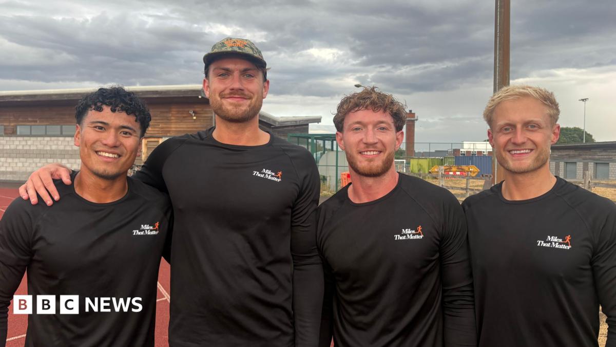 Four men standing on a running track. They are all wearing long-sleeve black tops with a white and orange Miles That Matter logo on the chest. The man on the left has black curly hair. The man next to him has brown facial hair and is wearing a camouflage cap. Next to him is a man with brown curly hair and facial hair. The man on the right has short, blond hair. The sky is cloudy and grey.