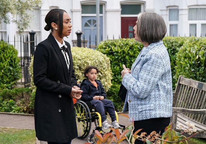 568588,TITLE:Eastenders Sheila and Chelsea chat in the Square, with Jordan in his wheelchair behind in a scene from EastEnders