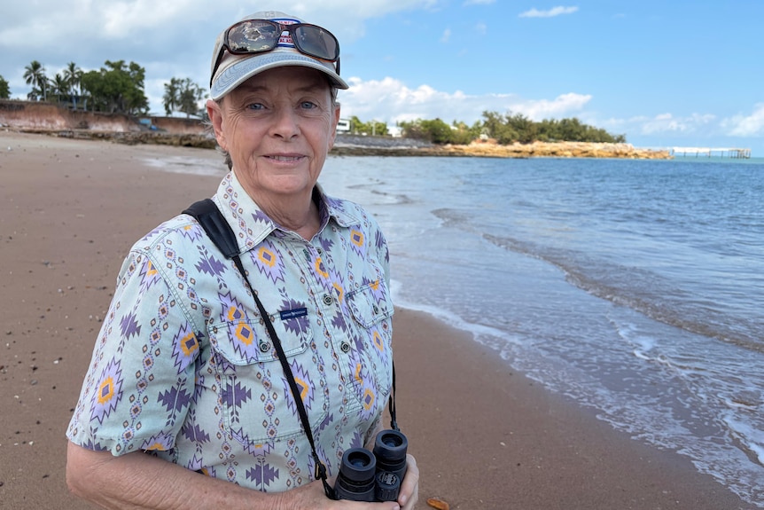A woman in a blue patterned collar shirt and cap, with binoculars in hand looking at camera as she stands on beach.