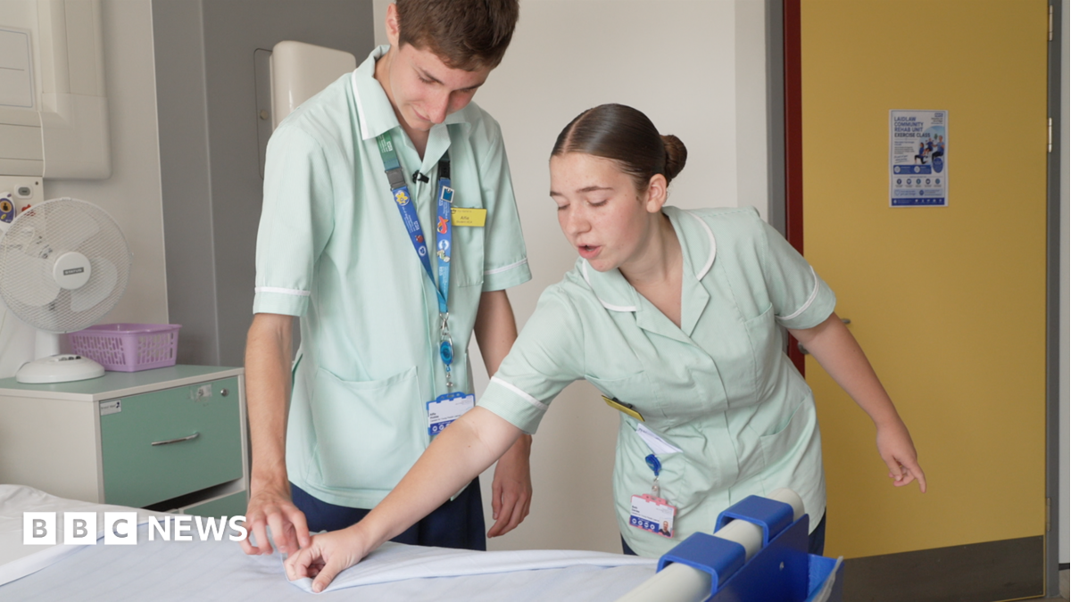Two hospital workers, a man and a woman, are pictured making a bed in a hospital room. They are wearing light green uniforms and lanyards.