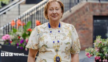 Rachel Nolan is smiling at the camera standing outdoors in front of flower beds and a staircase. She is wearing a patterned cream dress and a presidential gold and blue chain around her neck.
