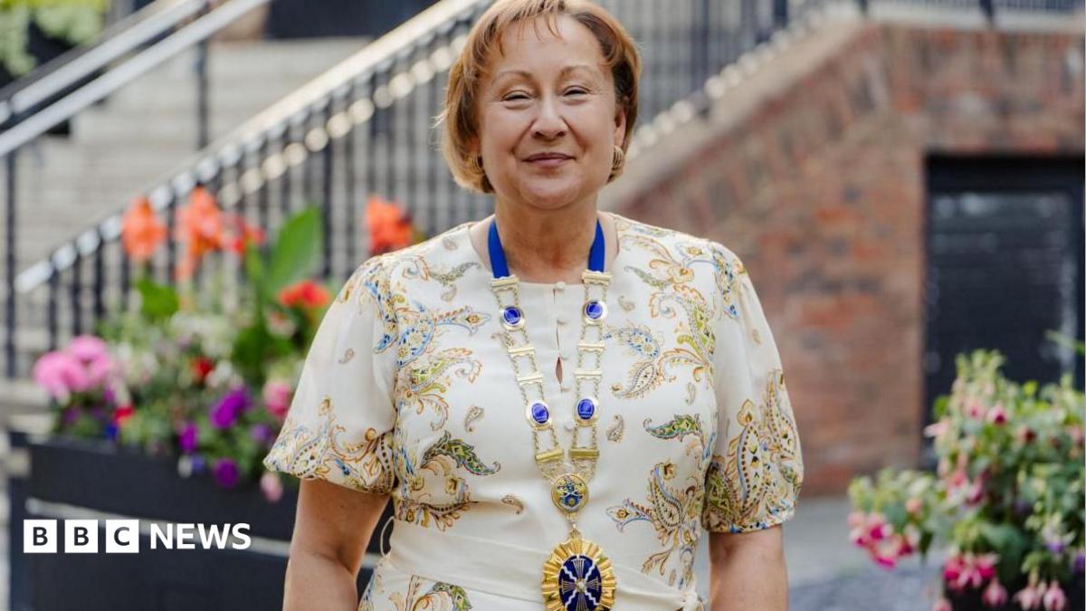 Rachel Nolan is smiling at the camera standing outdoors in front of flower beds and a staircase. She is wearing a patterned cream dress and a presidential gold and blue chain around her neck.